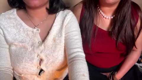 Video of two women in casual attire: one in a white textured cardigan with a necklace, the other in a red sleeveless top and a pearl necklace, seated side by side.