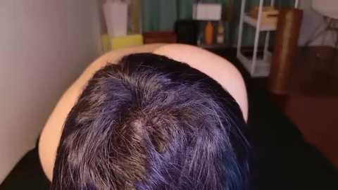 Video of a person with short, black hair bent over a table, seen from behind. Background shows a cluttered room with a white shelf, yellow box, and brown chair.