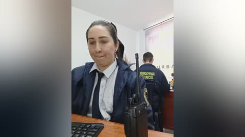 A video of a young woman with light skin and dark hair tied back, wearing a navy-blue police uniform and white shirt, seated at a desk. Background shows two officers in uniform.