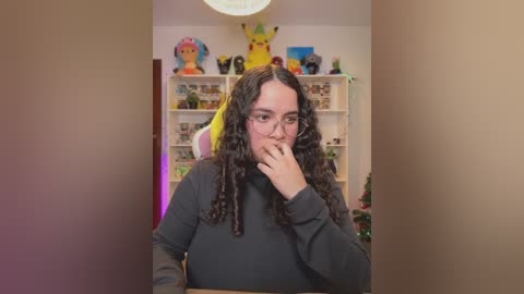 Video of a young woman with long, curly hair and glasses, wearing a dark sweater, standing in a brightly lit room with colorful Pok\u00e9mon decorations and shelves filled with books.