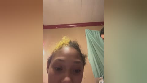Video of a young Black woman with curly, yellow-dyed hair, wearing a green shirt, in a room with beige walls and a white ceiling.