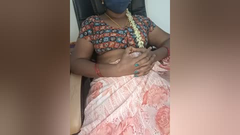 A video of a woman lying on a bed, wearing a colorful, patterned blouse, pink sari, and blue mask, with a red bangle on her wrist.