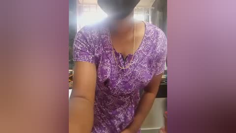 Video of a woman with medium brown skin, wearing a purple floral dress with a V-neckline, standing indoors in a dimly lit room with blurred background.