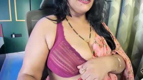 Video of a plus-sized woman with long black hair, wearing a deep purple lace bra and a pink floral sari, sitting in a modern room with green walls and floral curtains.
