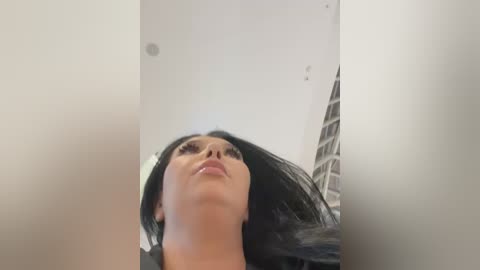 A low-angle video of a woman with long black hair, wearing makeup, standing in a modern indoor setting with white walls and a high ceiling.