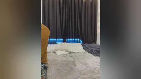Video of a dimly lit bedroom with a beige bed, a white pillow, dark curtains, and a blue light strip at the base, giving a cozy ambiance.