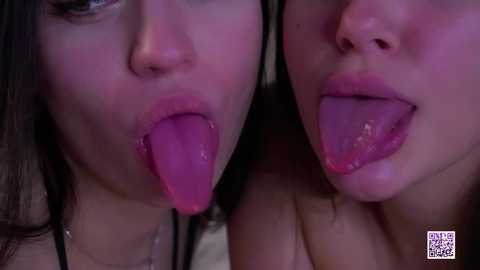 Video of two women with fair skin and dark hair, licking each other's tongues. Background is dimly lit, with a watermark in the bottom right corner.
