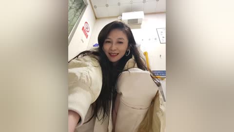 A video of an Asian woman with long black hair and fair skin, wearing a beige coat, smiling in an indoor setting with white walls, a sign, and a window.