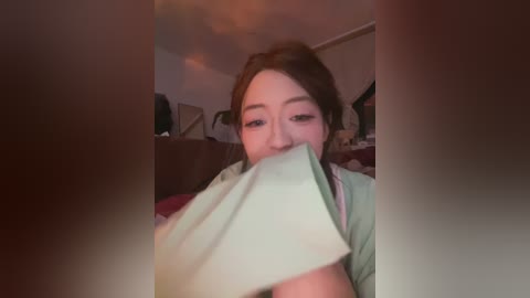 Video of a young Asian woman with light skin, brown hair, and minimal makeup, holding a piece of paper over her mouth. She's indoors, on a couch with a plush toy and a red blanket in the background.