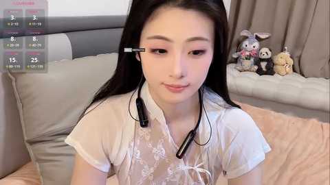 A video of an East Asian woman with long black hair, wearing a sheer white top, headphones, and a serene expression. Background includes a gray bed with stuffed animals, a white pillow, and beige curtains.