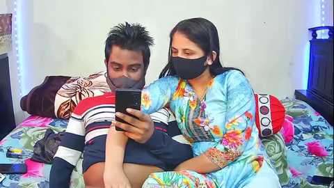 Video of a young South Asian couple in traditional outfits on a bed, using a smartphone; woman in floral saree, man in striped shirt, both masked.