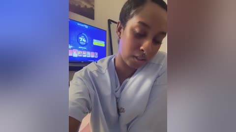 Video of a young woman with light brown skin and dark hair, wearing a light blue shirt, leaning forward. A TV screen in the background shows a weather forecast.