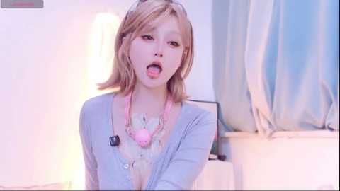 Media: A young, light-skinned girl with shoulder-length blonde hair, wearing a light blue cardigan, is playfully sticking out her tongue in a brightly lit room with white walls and a blue curtain.