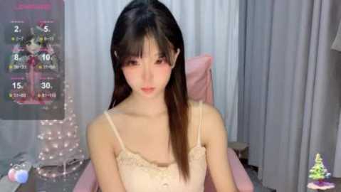 Video of a young East Asian woman with long black hair, wearing a pink spaghetti-strap top, seated indoors in front of a pink gaming chair.
