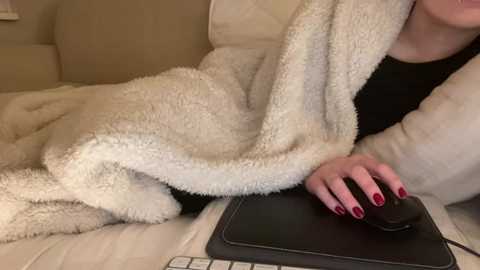 Video of a person, partially visible, wearing a fluffy white blanket, resting on a beige couch, with a black mouse and keyboard on their lap.