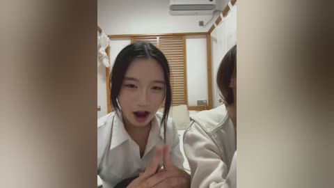 Video of a young Asian woman with long black hair, wearing a white shirt, looking surprised and open-mouthed, partially obscured by hands in the foreground. Background features wooden paneling and an air conditioner.