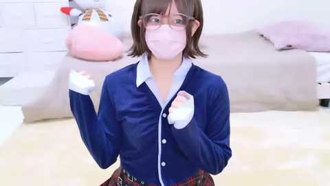 Video of a young woman with short brown hair, wearing glasses, a blue blouse, and a face mask, playing a virtual reality game in a brightly lit, minimalist room.
