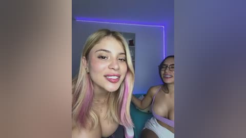 Video of two young women with fair skin, blonde hair, and pink streaks, smiling; one topless, the other wearing a white top. Background shows a dimly lit room with blue LED strip lights.