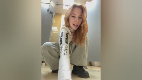 Video of a smiling, light-skinned, red-haired woman with long hair in a grey and white Star Wars-themed hoodie and grey sweatpants, squatting in a hallway with beige tiled floor.