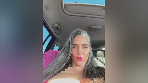 A video of a woman with long, gray-streaked hair, wearing a pink top, sitting in a car with a light interior, making a kissy face.