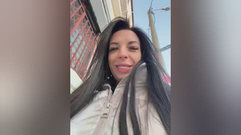 Video of a smiling, dark-haired woman with long hair, wearing a light-colored jacket, standing in an urban setting with red metal railings and a blue sky.