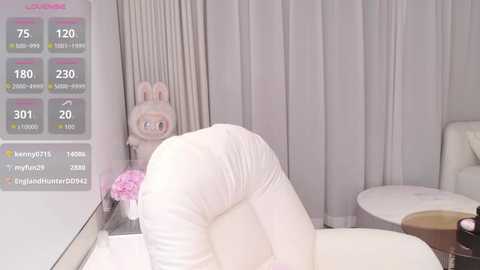 A video of a plush, cartoonish bunny with large ears, wearing white pajamas, posing provocatively on a white bed in a softly lit, modern room with grey curtains.