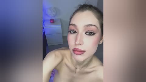 A video of a fair-skinned, light-haired woman with pink eyeshadow, red lipstick, and a bare upper body, standing against a white wall with a blue-lit vase and pink flower in the background.