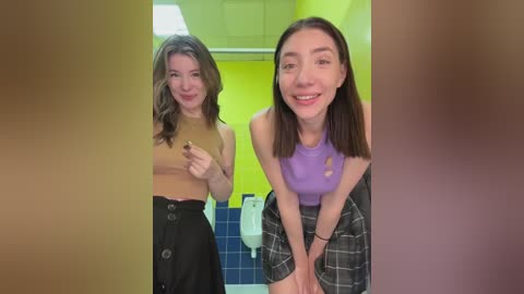 Media: Video of two women in a public restroom: one with a cropped top and black skirt, the other with a purple top and plaid skirt, both smiling. Background shows yellow walls and blue tiles.