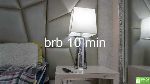 Media: Video of a modern bedroom with a white bed, geometrically patterned headboard, and a white nightstand holding a silver lamp and remote control. Text overlays \"brc 10 min.\