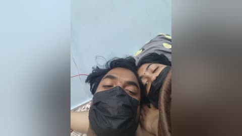 Video of a South Asian man with medium skin tone and black hair, wearing a black mask and gray shirt, lying on a bed with a brown blanket, with a blurred, possibly distressed young child in the background.