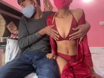 Video of a man wearing a mask, blue shirt, and jeans, embracing a woman in a red bikini and matching robe, both seated on a bed with pink walls in the background.