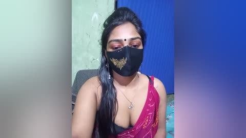 Video of a South Asian woman with medium-brown skin and long black hair, wearing a black mask, red sari, and a black bra. She sits in a chair against a blue and green wall, with a subtle blue gradient overlay.