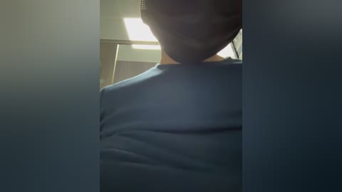 Media: Video of a person wearing a dark mask, standing in a dimly lit hallway with a bright ceiling light. The person is wearing a dark blue shirt.