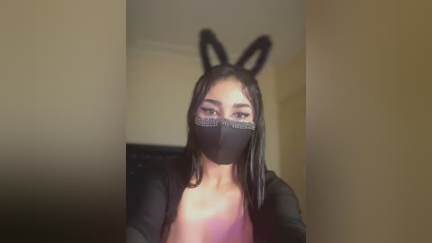 Video of a young woman with long dark hair, wearing a black face mask, bunny ears headband, and a sheer black top, standing indoors against a beige wall.