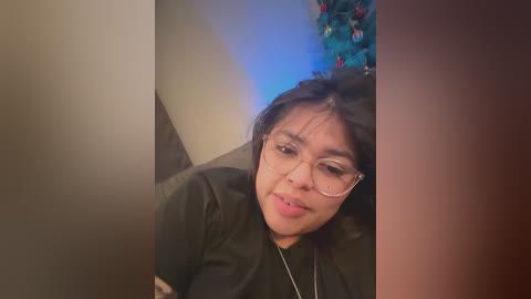 Video of a young Asian woman with glasses, light skin, and black hair, smiling, wearing a black jacket, seated indoors against a blurred, festive background with a blue light and a decorated Christmas tree.