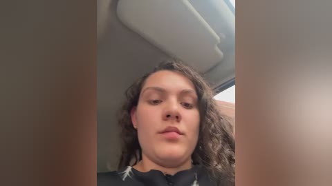Video of a young, light-skinned person with curly brown hair, wearing a black t-shirt, sitting in a car with a beige interior.