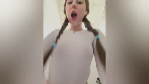 Video of a young Caucasian woman with fair skin and light brown hair in twin braids, wearing a tight white top, looking surprised or shocked, taken from a low angle.