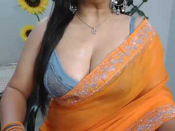 Video of a South Asian woman with medium skin tone, wearing an orange sari with a blue lace bra visible, adorned with silver jewelry, in a room with sunflower decor.