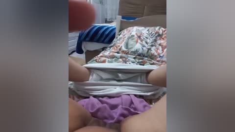 A video of a person in a light-colored, floral-patterned shirt and purple shorts, lying on a bed with beige and blue bedding, blurred background.