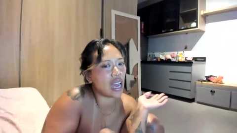 Video of a topless Asian woman with short black hair, medium skin tone, and visible tattoos, crying and holding her mouth, in a modern, dimly-lit room with kitchen cabinets and a counter.