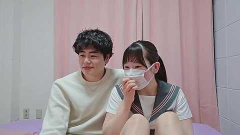 Video of two Asian individuals wearing face masks, sitting on a bed in a clinical room with pale pink curtains and white walls.