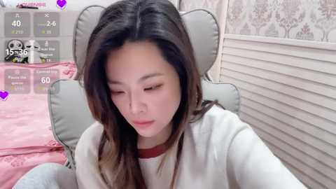 A video of an East Asian woman with long, dark hair, wearing a white sweater, sitting on a gray chair in a pink room with floral wallpaper.