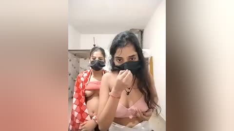 Video of two women in a bathroom. One wears a red-patterned saree, the other a pink bra, both with black masks. The room has beige walls and a tiled floor.