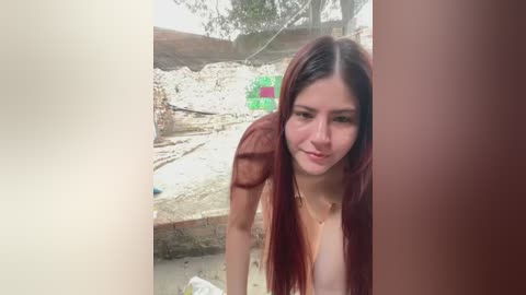 Video of a young, fair-skinned woman with long, straight auburn hair, partially covered by a sheer, red scarf. She's topless, smiling softly. Background features a construction site with exposed earth, green and red plastic barriers, and trees.