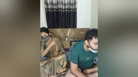 Video of an Indian woman in a golden sari and a man in a green T-shirt, both masked, lying on a bed with patterned sheets, in a dimly lit room with dark curtains.