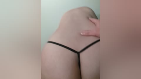 A blurred video of a person wearing a black thong, with a hand on their buttocks. The background is a plain, light-colored wall.