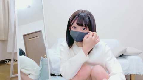 Video of an Asian woman with straight black hair, wearing a gray face mask, white sweater, and light blue overalls, sitting on a bed in a minimalist, white room.