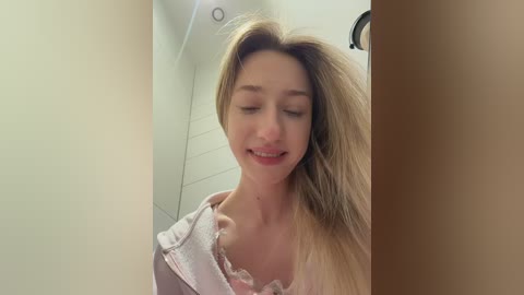 Video of a young Caucasian woman with long blonde hair, wearing a light pink shirt, smiling, standing in a bathroom with white tiled walls.
