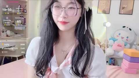 A video of a young Asian woman with long black hair, glasses, and a white shirt tied at the waist, posing indoors with plush toys and framed artwork in the background.