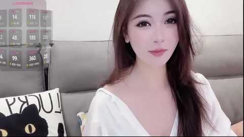 Video of a young East Asian woman with long brown hair, fair skin, and brown eyes, wearing a white off-shoulder blouse. She sits on a gray couch, with a black and white pillow featuring \"MAD!\" in the background.